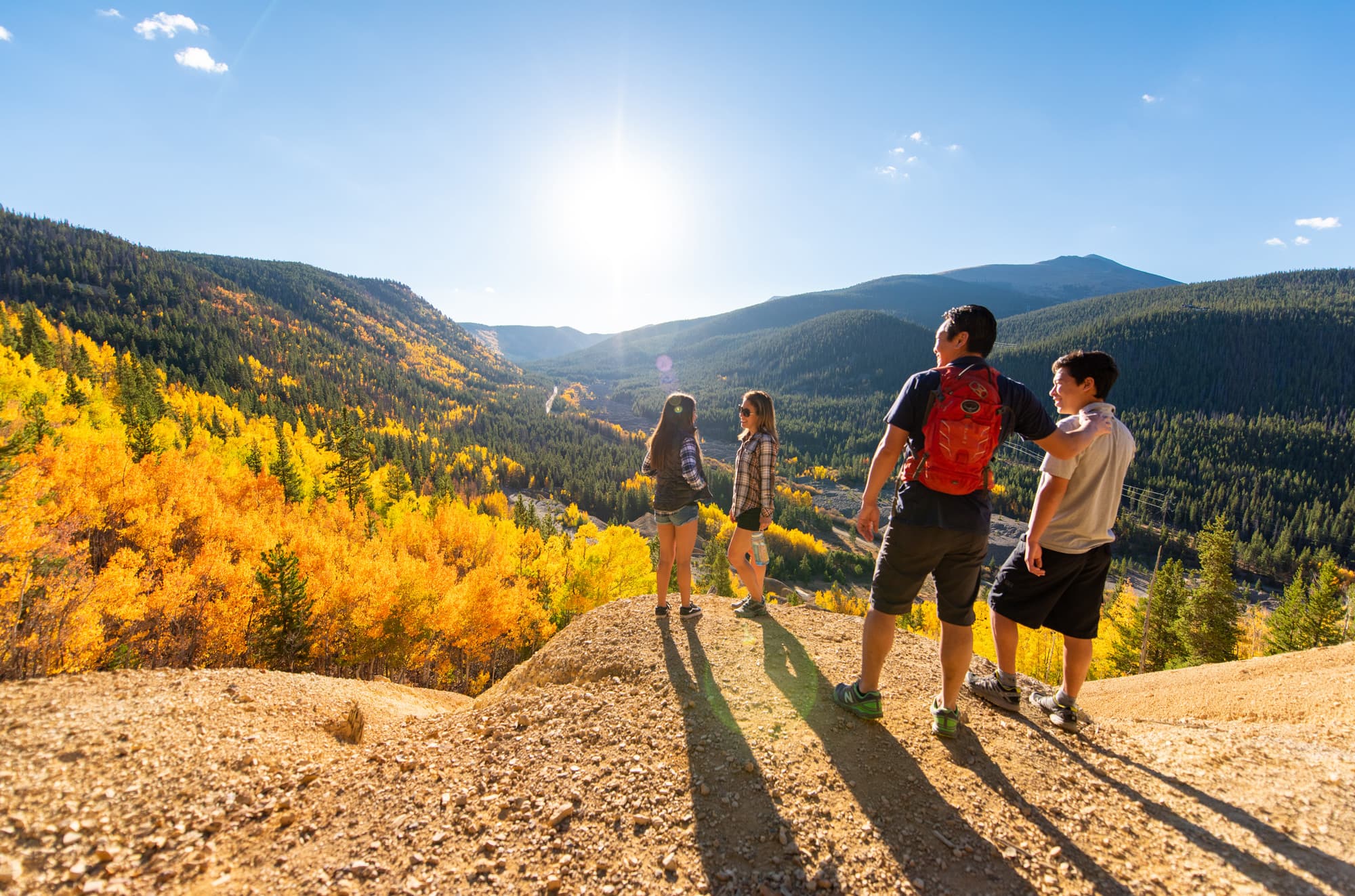 Family looking out over a valley on a sunny day. Yellow trees are seen throughout the mountains.