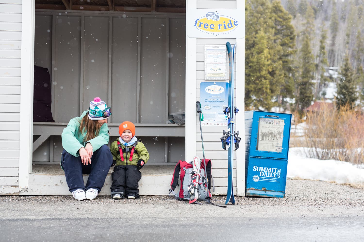 Parent and child sitting beside ski gear on a snowy day.